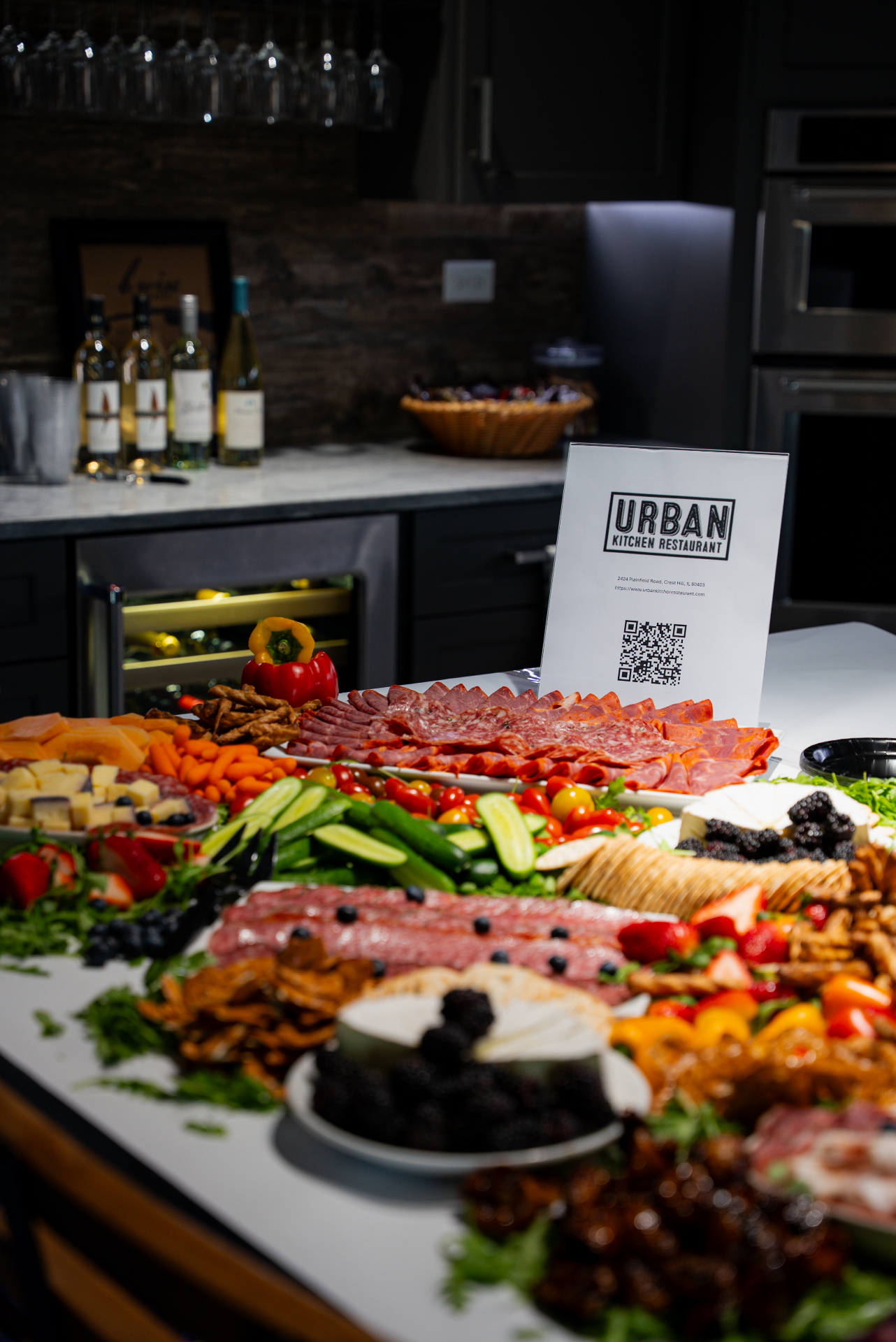 A large Urban Kitchen catering spread of charcuterie, cheeses, fresh vegetables, fruit, and wine on a kitchen island